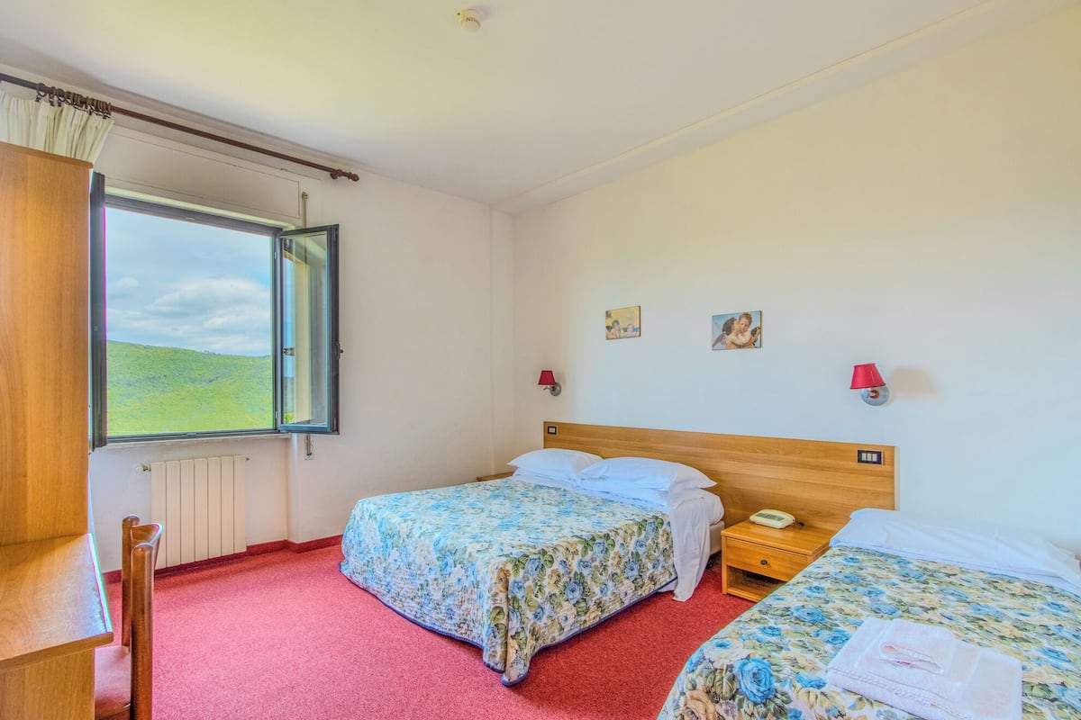 Une chambre d’hôtel avec deux lits, une tête de lit en bois, une fenêtre donnant sur un paysage verdoyant et une lampe rouge fixée au mur.