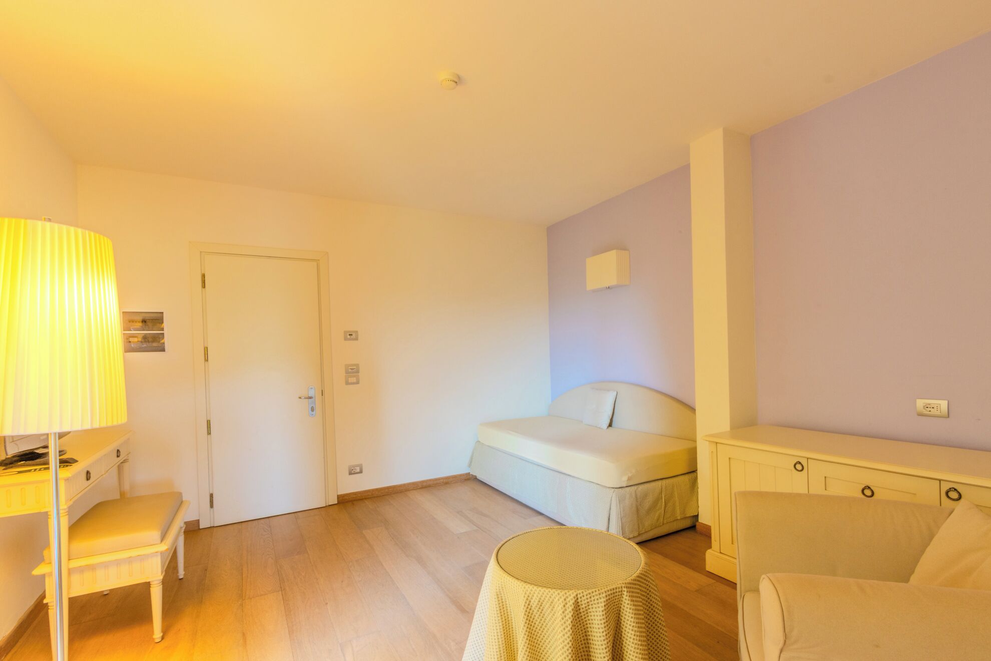 A room with a bed, a desk with a lamp, a chair, a small round table, and a cabinet.