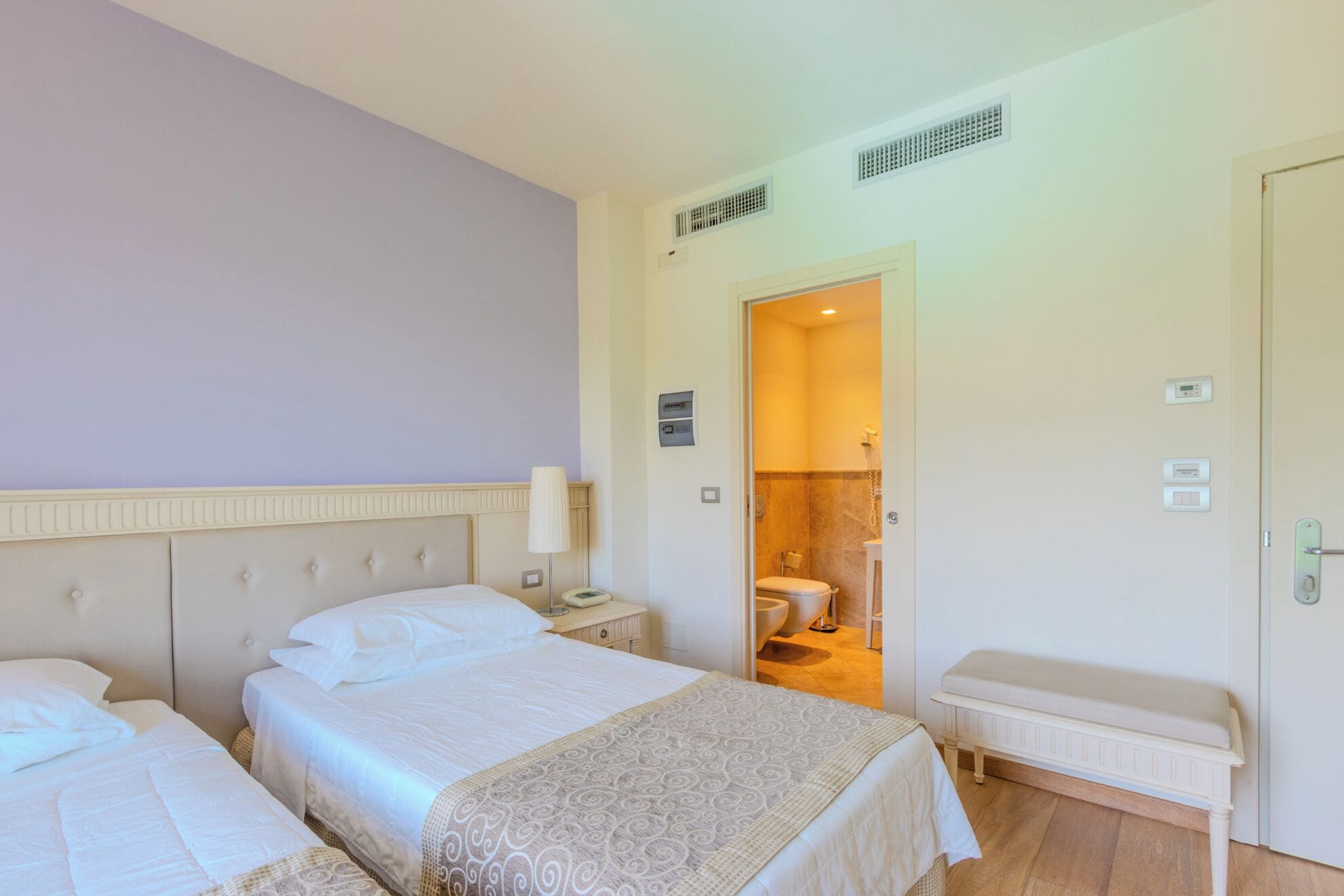 A hotel room with two beds, a bedside table with a lamp, a bench, a bathroom visible through an open door, and a thermostat on the wall.