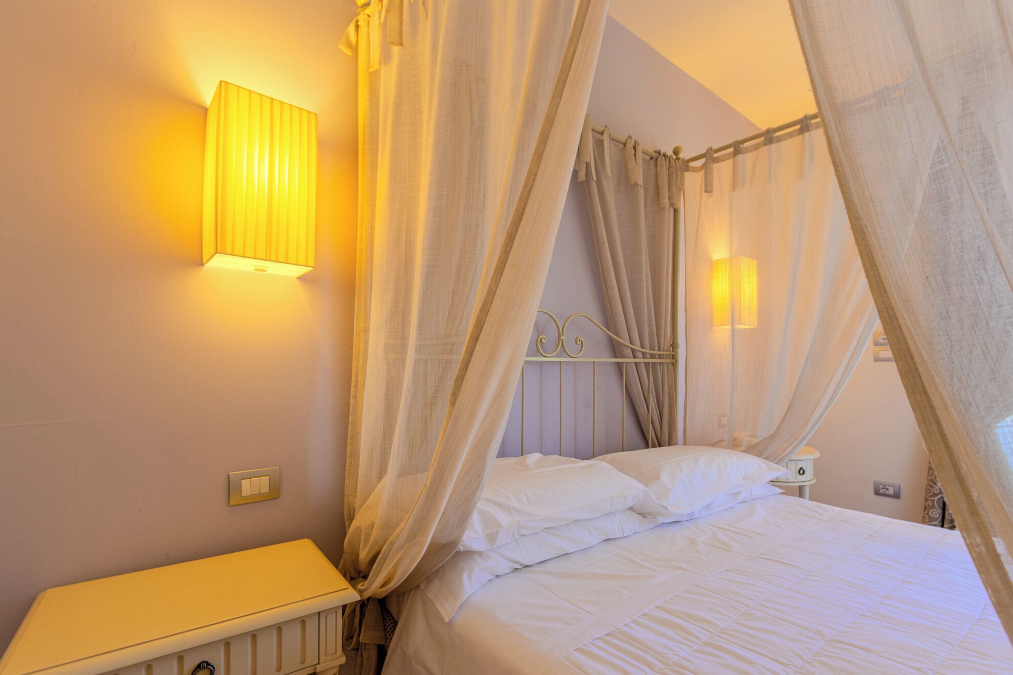 Ein Bett mit weißer Bettwäsche, ein Nachttisch, eine Wandleuchte und ein Vorhang.