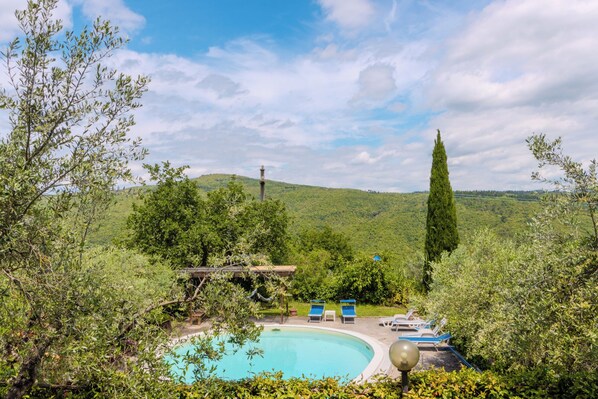 Una piscina rodeada de abundante vegetación, un pérgola de madera y una vista despejada de colinas distantes.