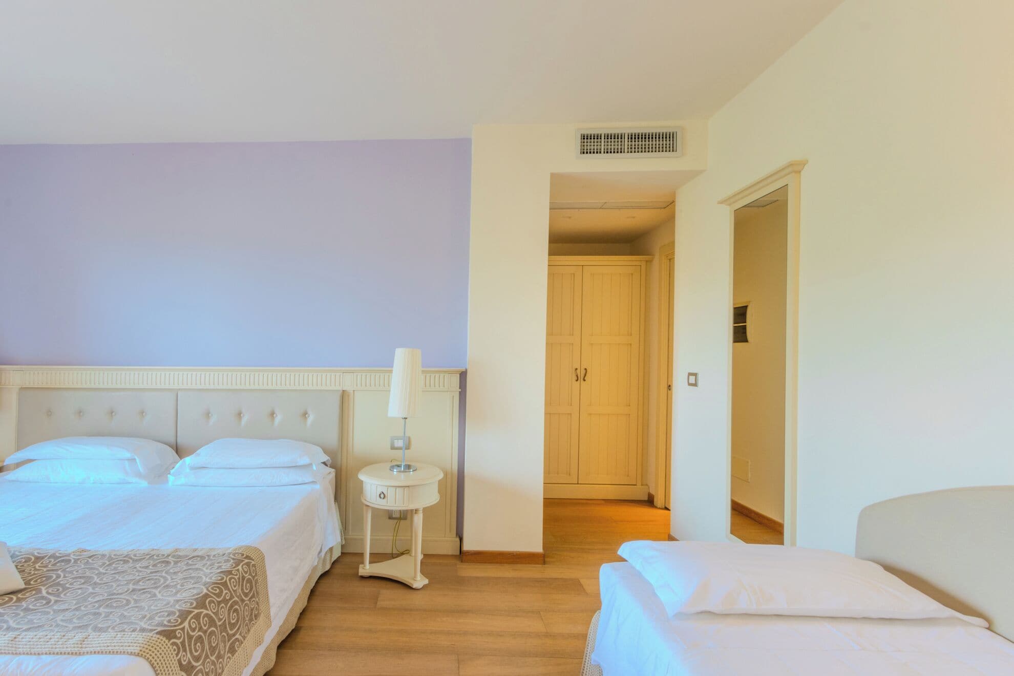 A hotel room with two beds, a bedside table with a lamp, a wardrobe, and a door leading to another room.