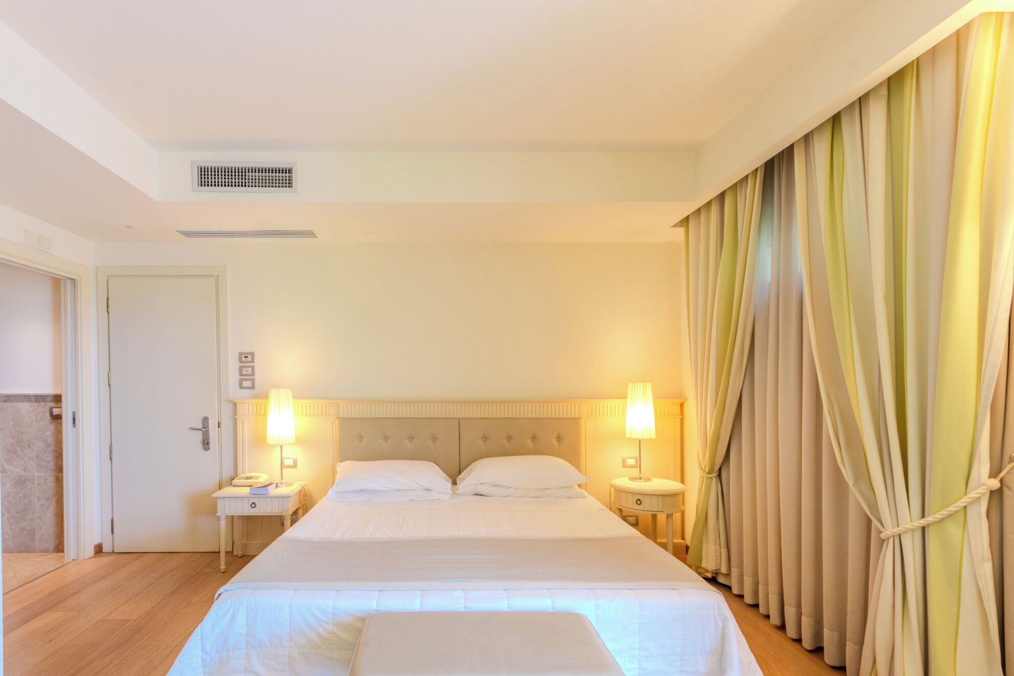 Une chambre d’hôtel avec un grand lit, deux tables de chevet, une petite table, une lampe et un rideau.