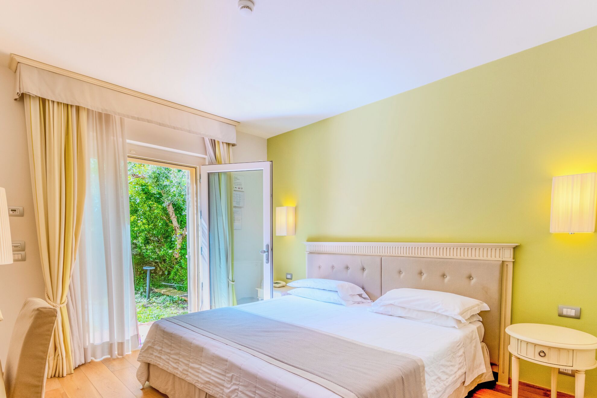 Ein Schlafzimmer mit Bett, Nachttisch, Lampe und Blick auf grüne Vegetation.