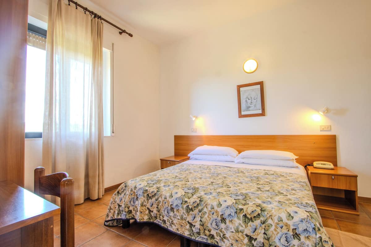 Une chambre d’hôtel avec un lit en bois, deux tables de chevet, une petite table, une chaise, une fenêtre avec des rideaux et un luminaire fixé au mur.