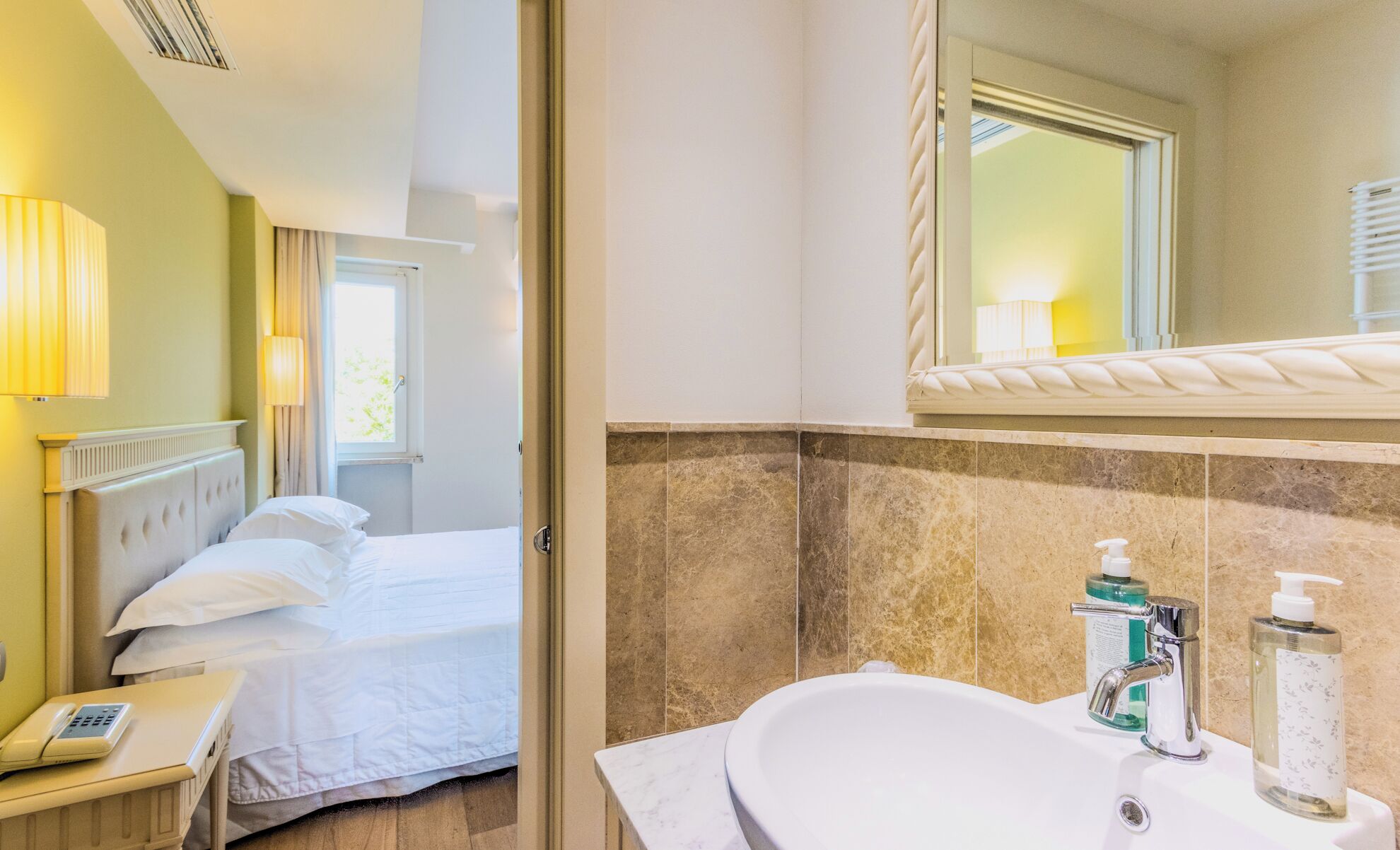 A hotel room with a bed, bedside phone, and a bathroom with a sink, mirror, and toiletries.