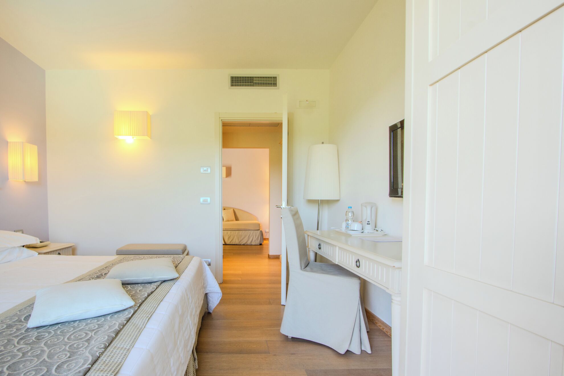 A hotel room with a bed, bedside table, lamp, and a view into the adjacent room.