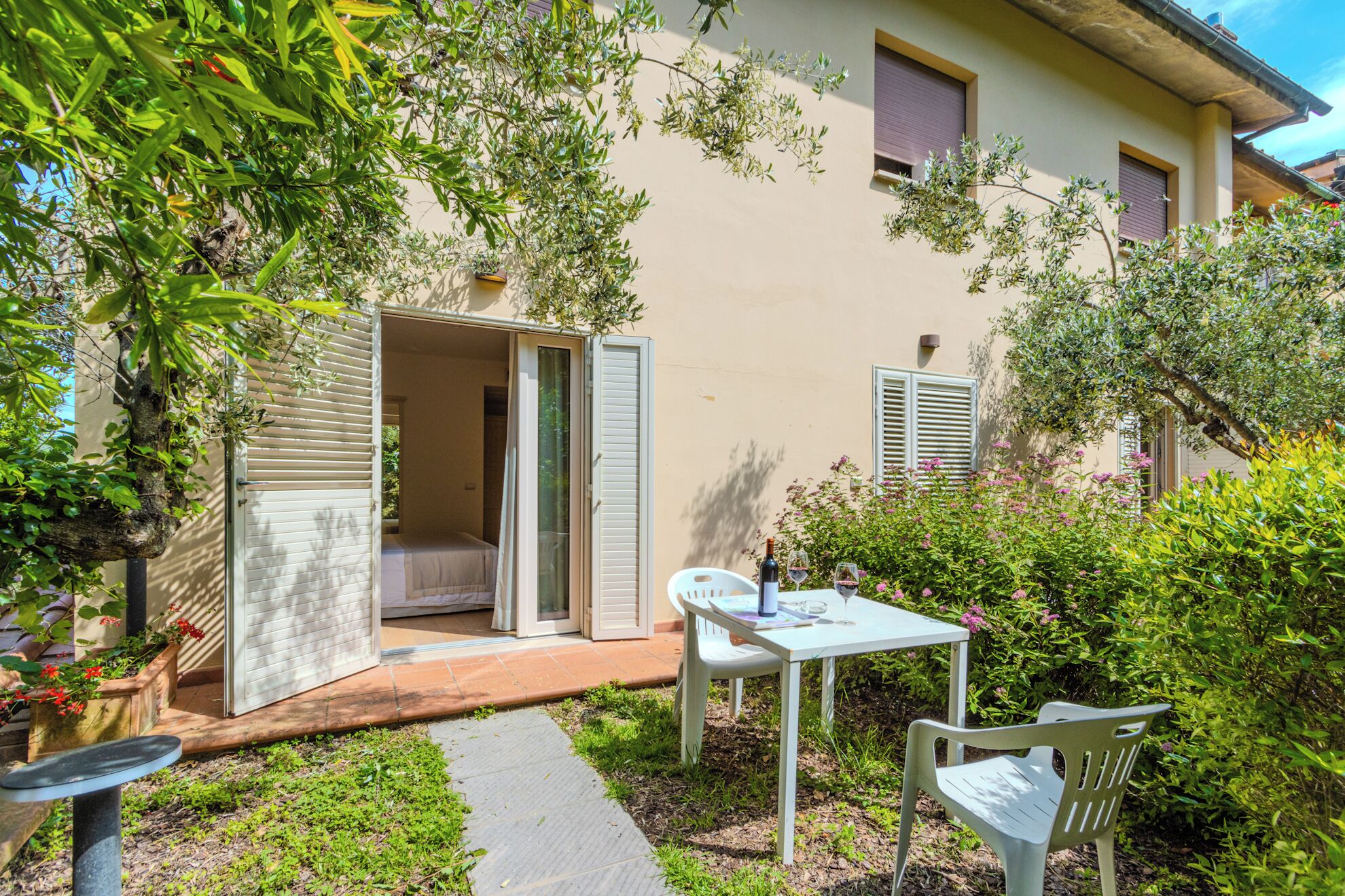 A small outdoor patio with a table and chairs, a door leading to a room, and surrounding greenery.