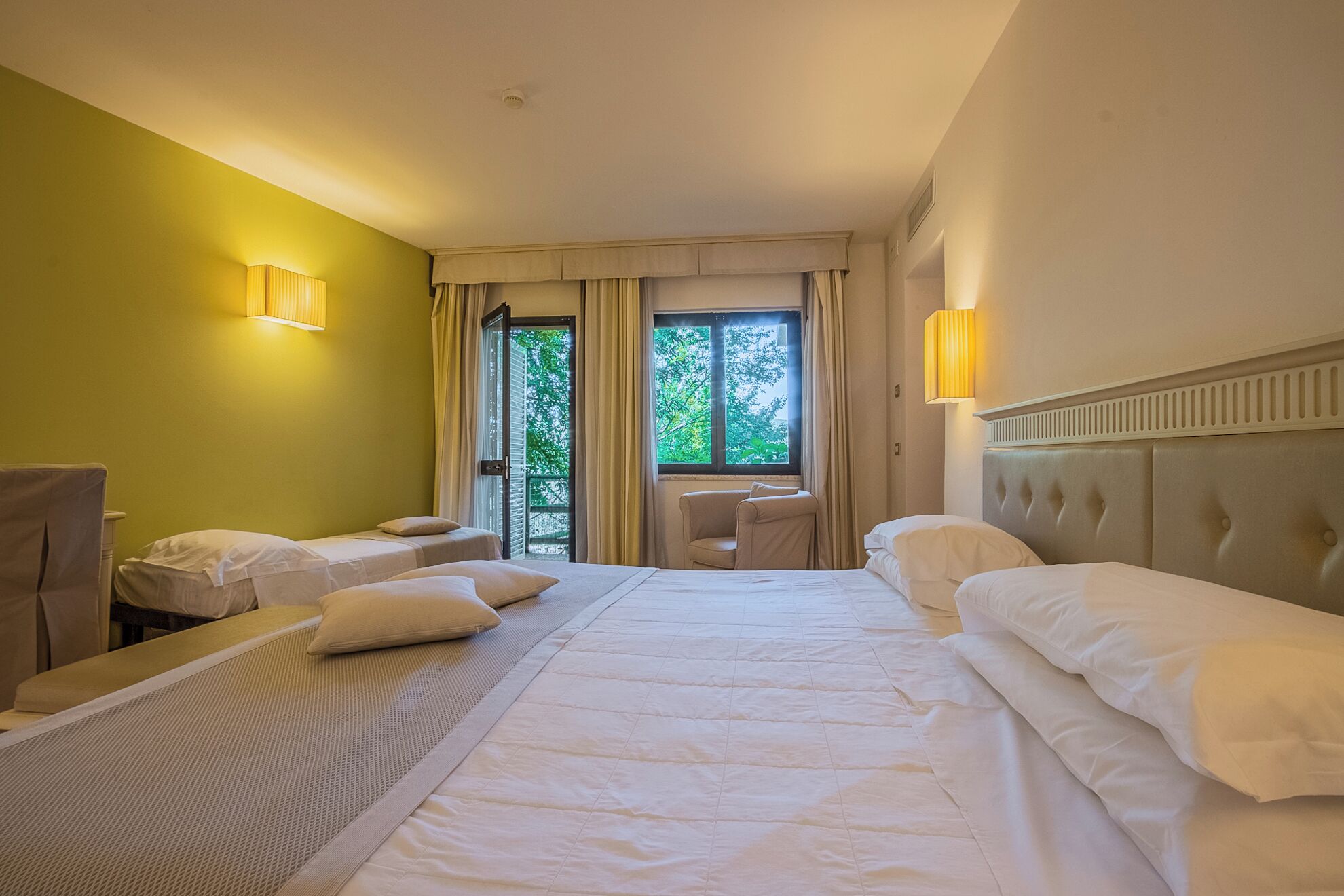 Une chambre d’hôtel comprenant un lit, deux tables de chevet, une chaise, une fenêtre avec des rideaux et une lampe fixée au mur.