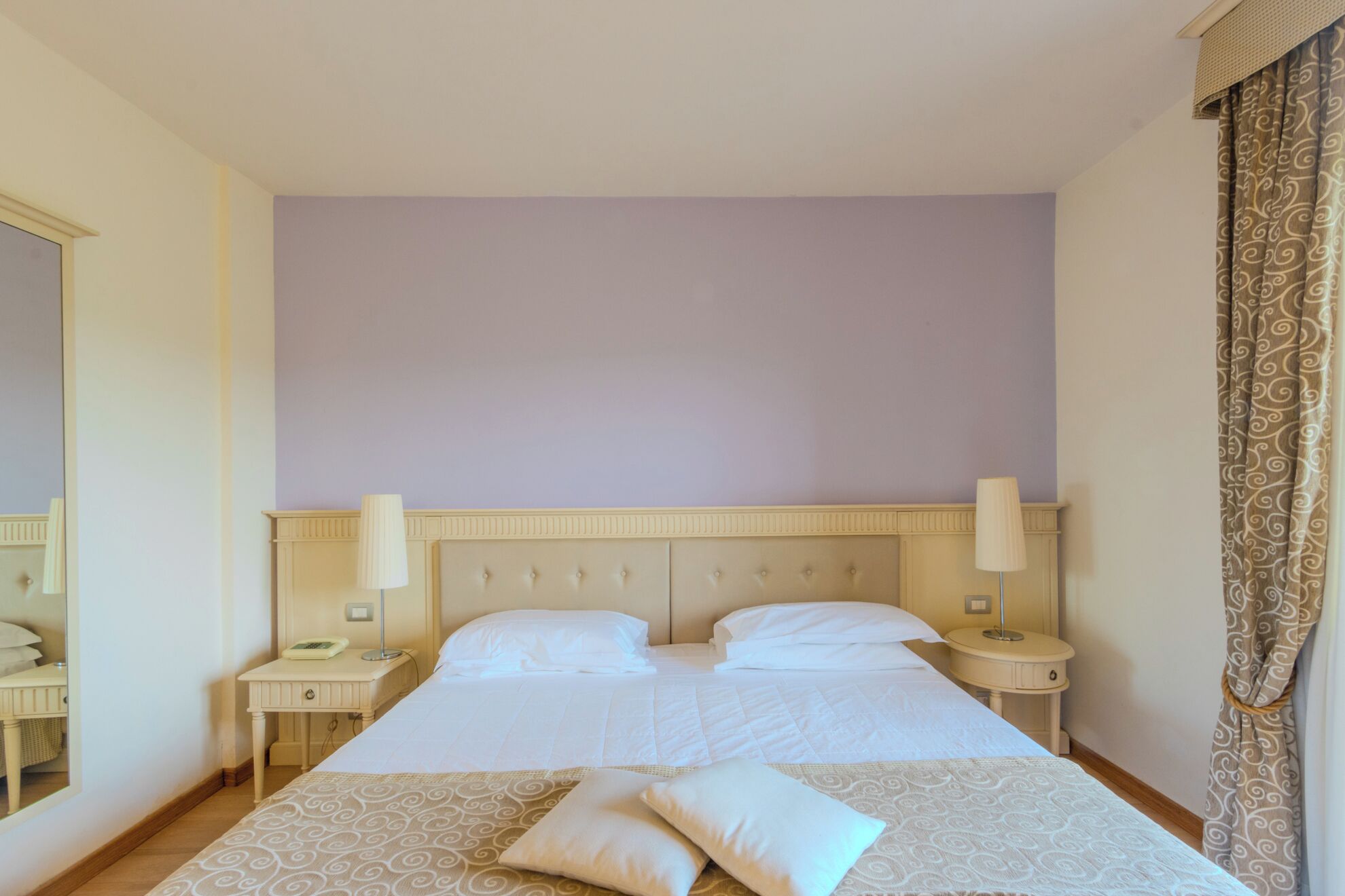 Une chambre d’hôtel avec un grand lit, deux tables de chevet, un miroir et une fenêtre avec des rideaux.