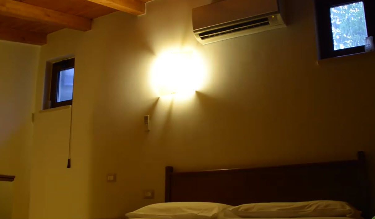 Une chambre avec un lit, une lampe fixée au mur, un appareil de climatisation et une petite fenêtre.