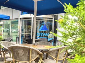 An outdoor seating area with wicker chairs and a table, a blue awning, and a glass door with signage. - Siyav Hotel (Ankara)