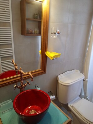 Un baño con un lavabo rojo, un grifo de madera, un espejo, un inodoro y un estante con artículos de aseo.