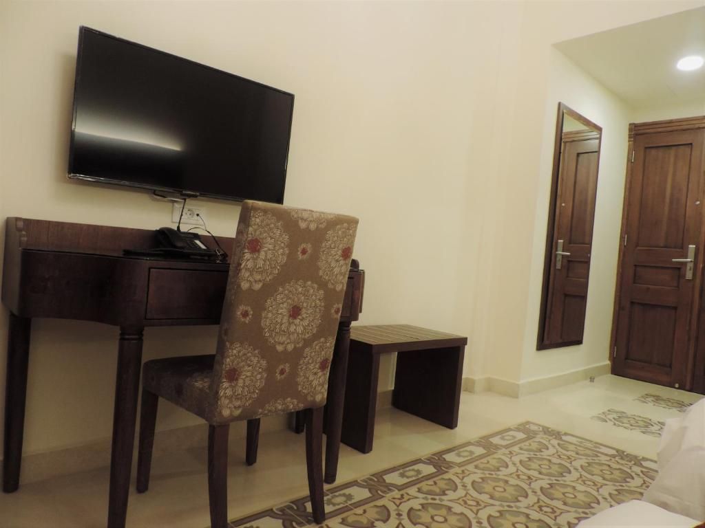 A room with a flat-screen TV mounted on the wall, a wooden desk, a patterned chair, a small wooden stool, and a patterned rug on the floor.
