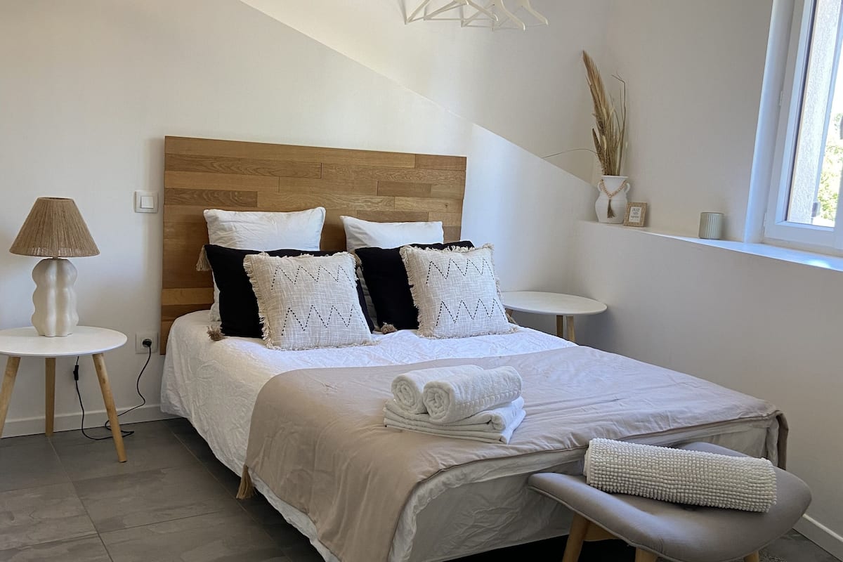 A bedroom with a wooden headboard, a bed with white and black bedding, a bedside lamp, a small table, a window with a view, and a decorative branch on the wall.