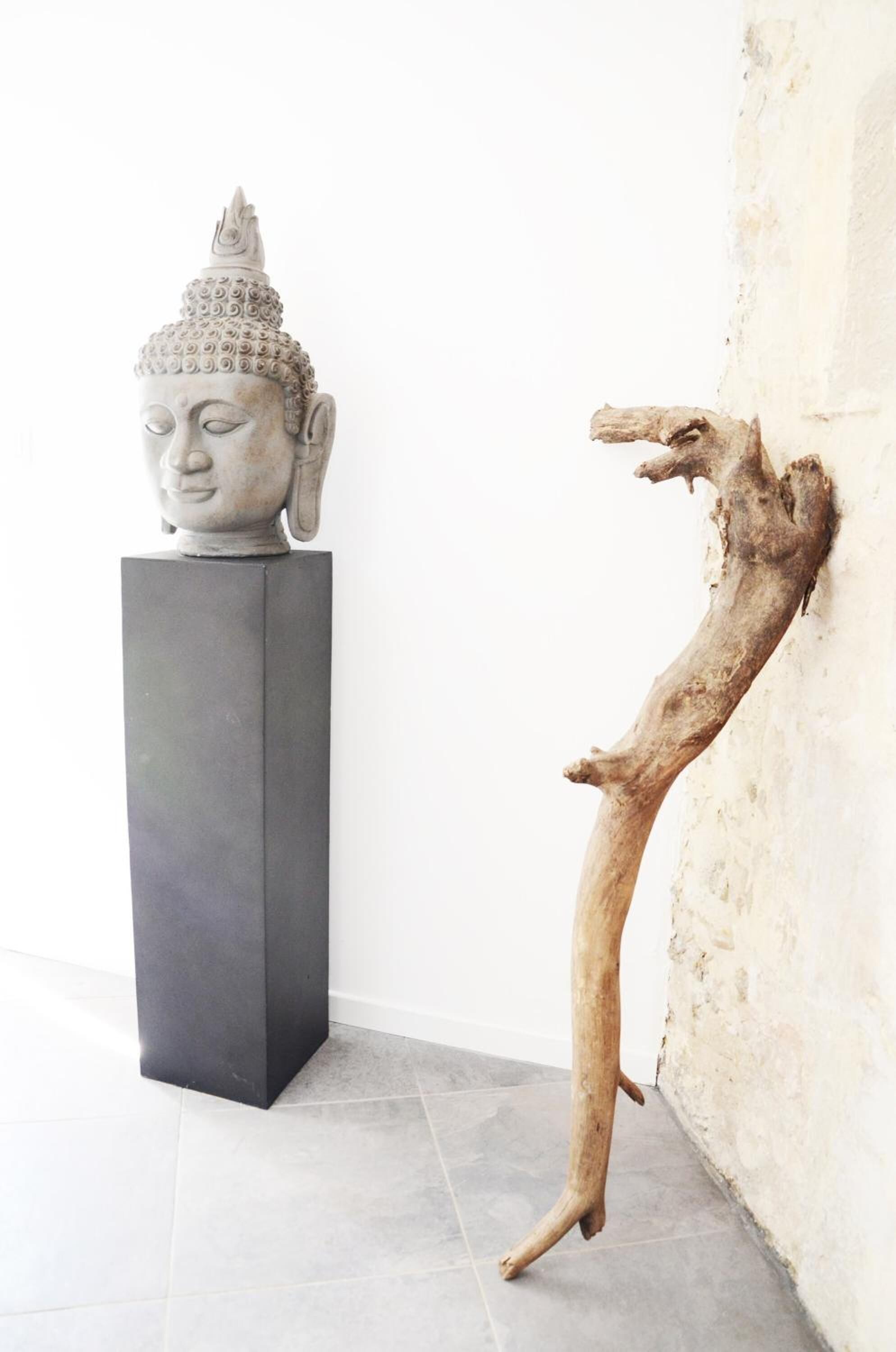A sculpture of a Buddha head on a pedestal, a wooden branch leaning against a wall, and a tiled floor.