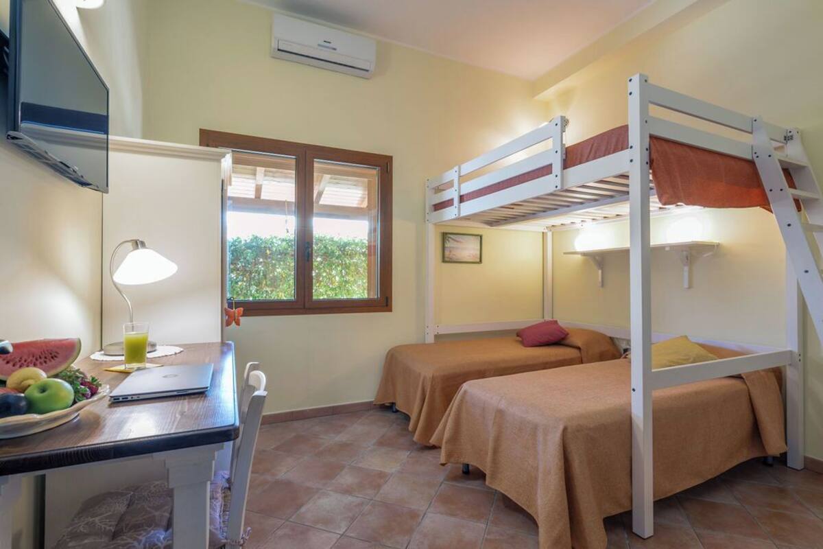 Een kamer met twee stapelbedden, een bureau met een laptop, een lamp en een raam met jaloezieën.