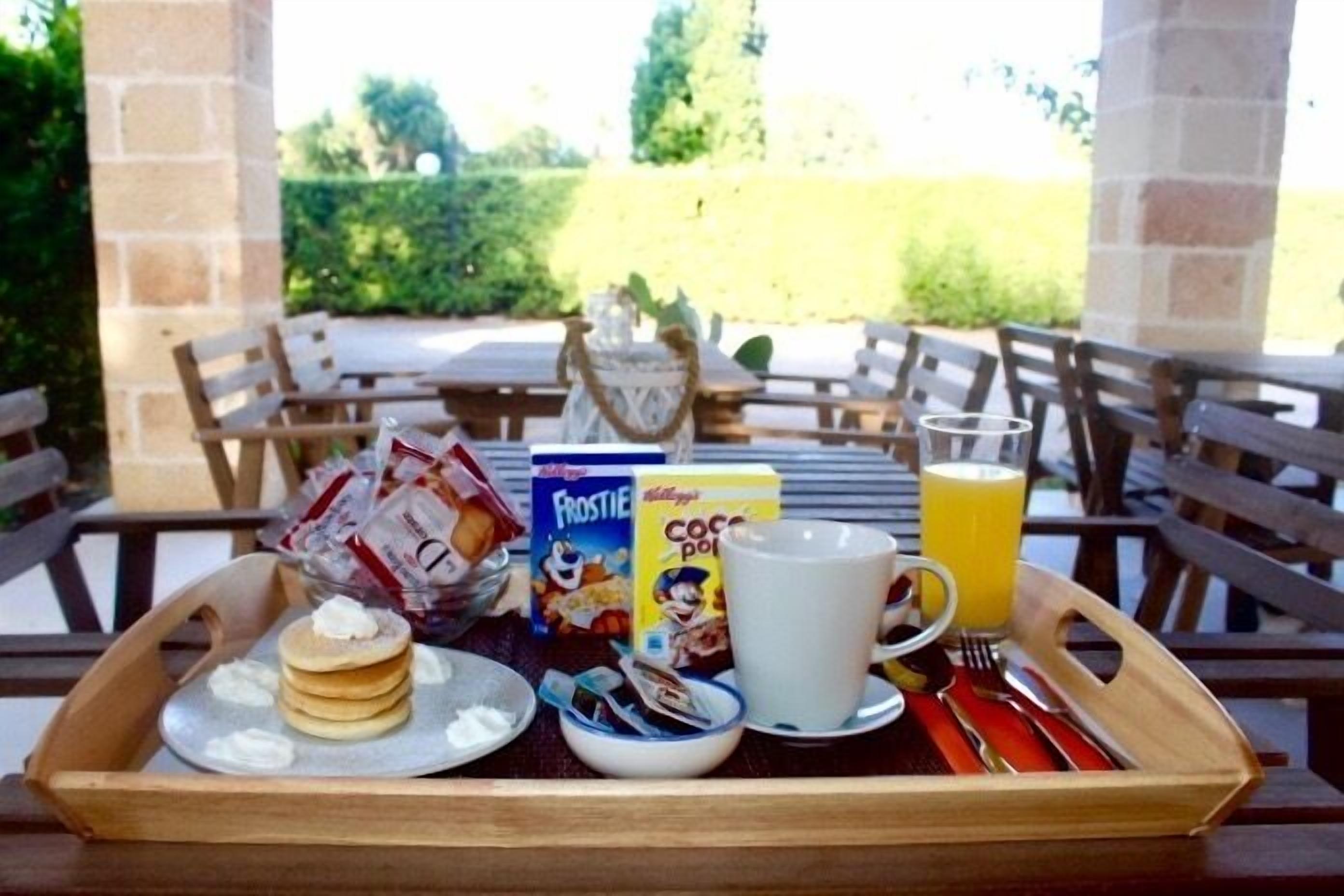 Ein Frühstücksbrett mit Pfannkuchen, Müsli, einer Tasse und einem Glas Orangensaft auf einem Holztisch im Freien.