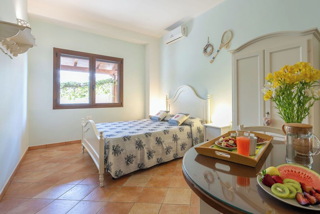 A bedroom with a bed, a bedside table with a tray of fruit, a pitcher, and a window with a view of greenery.