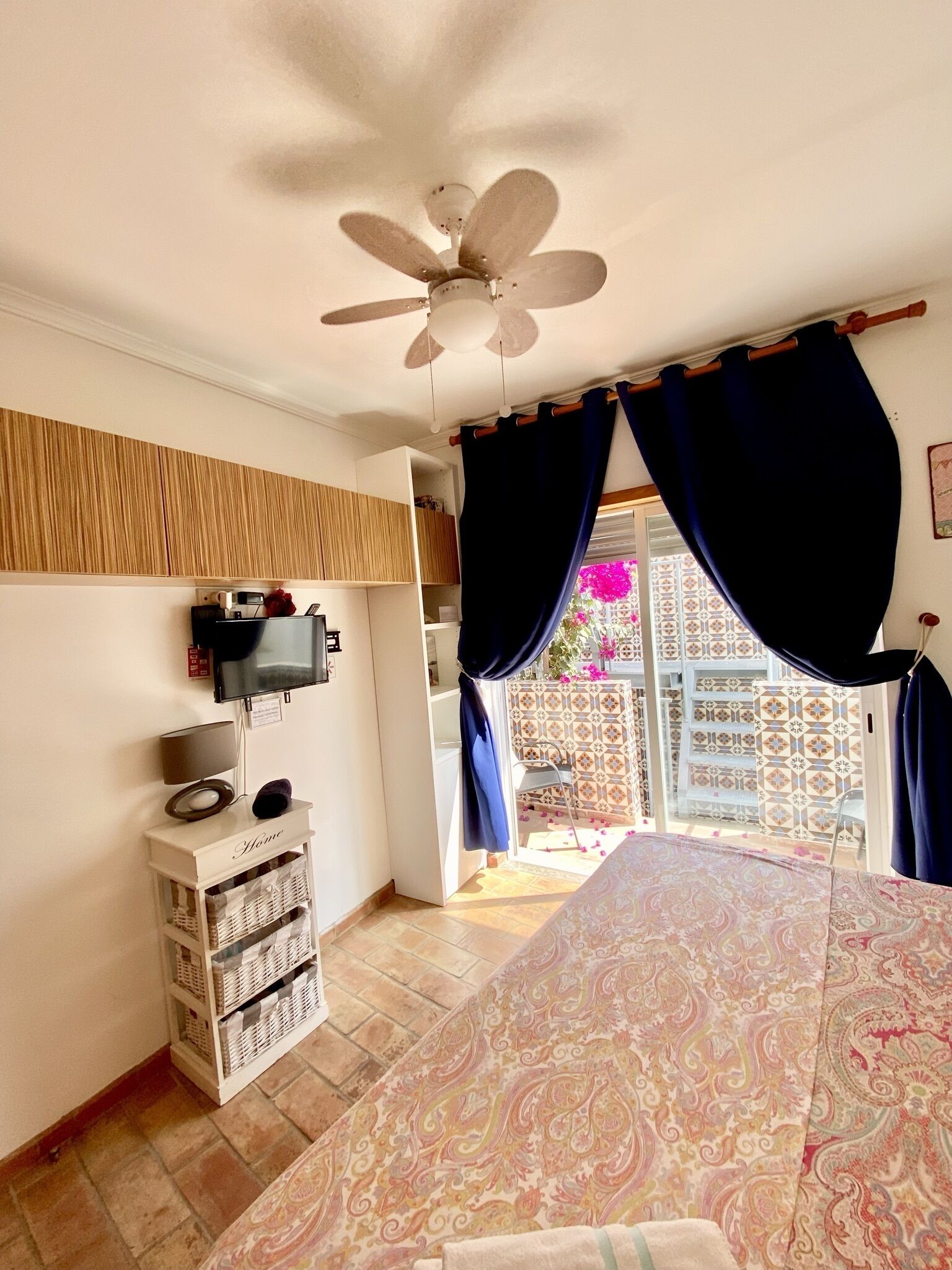 A bedroom with a ceiling fan, a TV mounted on the wall, a bedside table with a lamp, and a patterned bedspread.