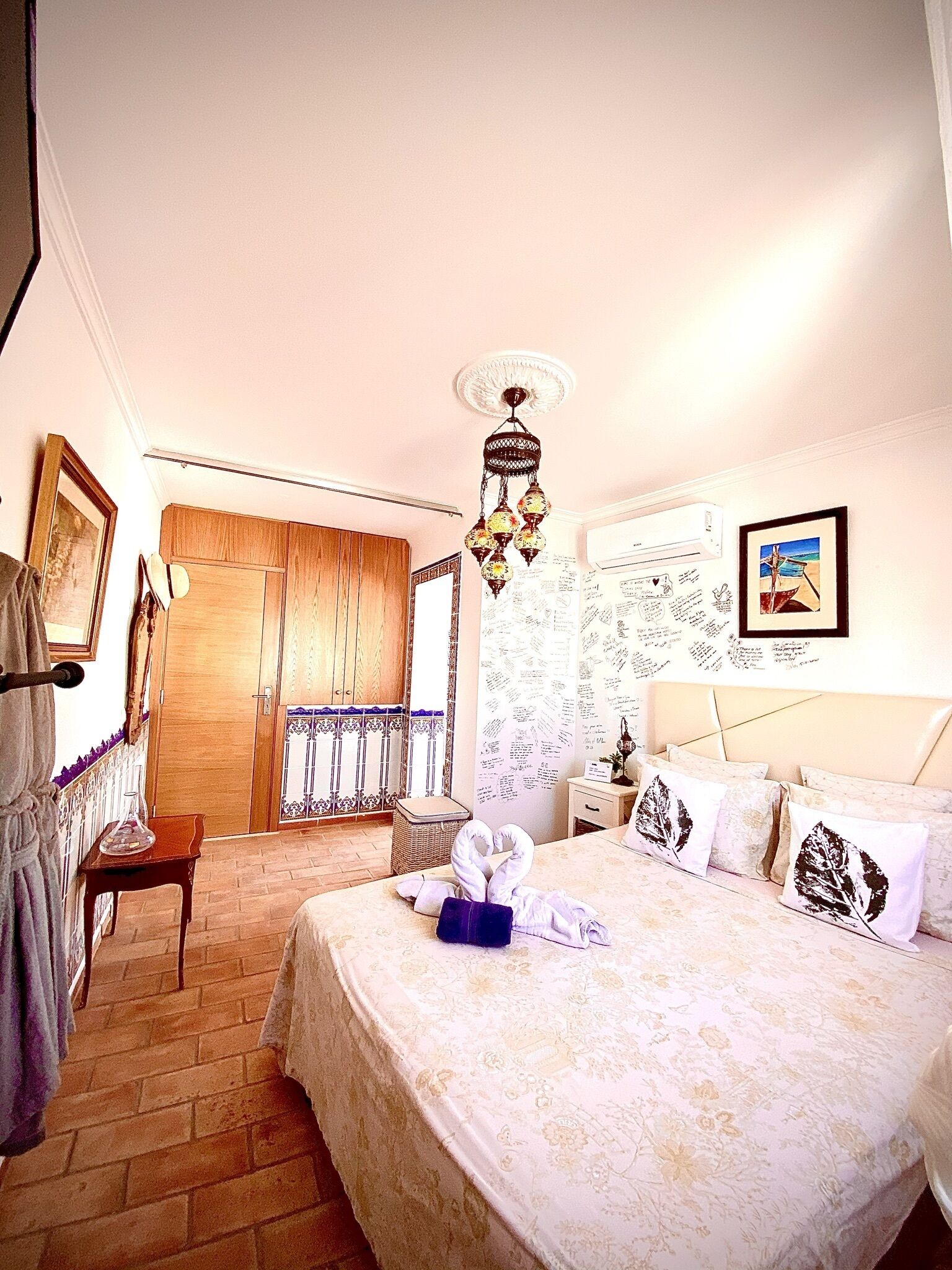 Hotelkamer met een bed, een houten garderobe, een tafeltje met een vaas, een decoratieve hanglamp en een muur met een lijst erin.