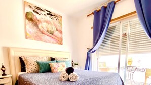 A bedroom with a large bed, a nightstand, a painting on the wall, and a view of the outside balcony. - Zenit Guest House (Olhao)