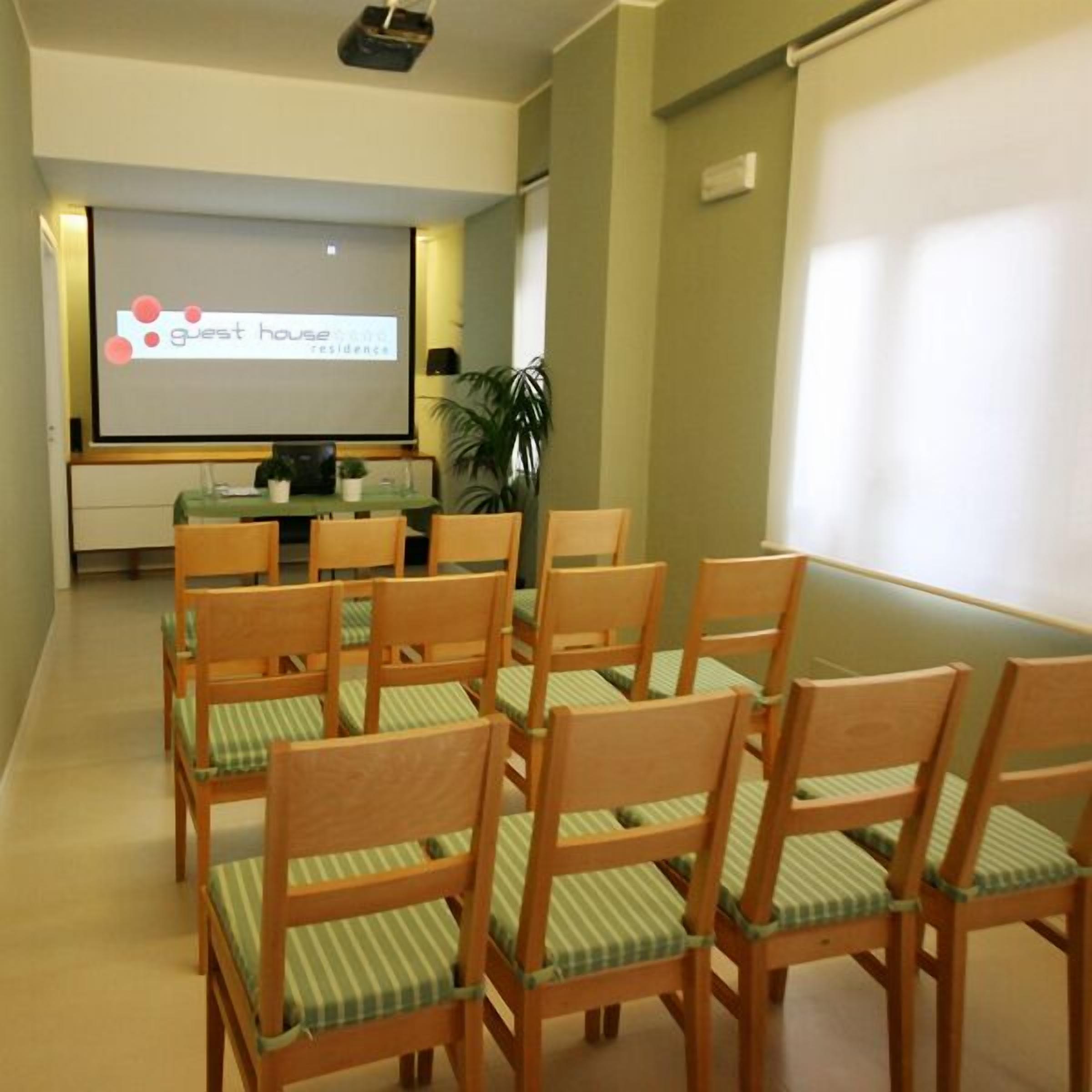 A conference room with a projector screen displaying "guest house residence" and several wooden chairs with green cushions.