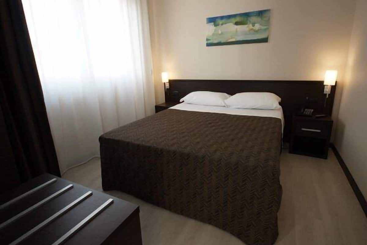 Une chambre d’hôtel avec un grand lit, deux tables de chevet avec des lampes, un tableau au mur et une fenêtre avec des rideaux.