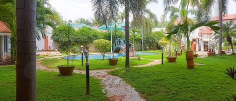 A lush garden with a pathway leading to a pool area, surrounded by palm trees and potted plants.