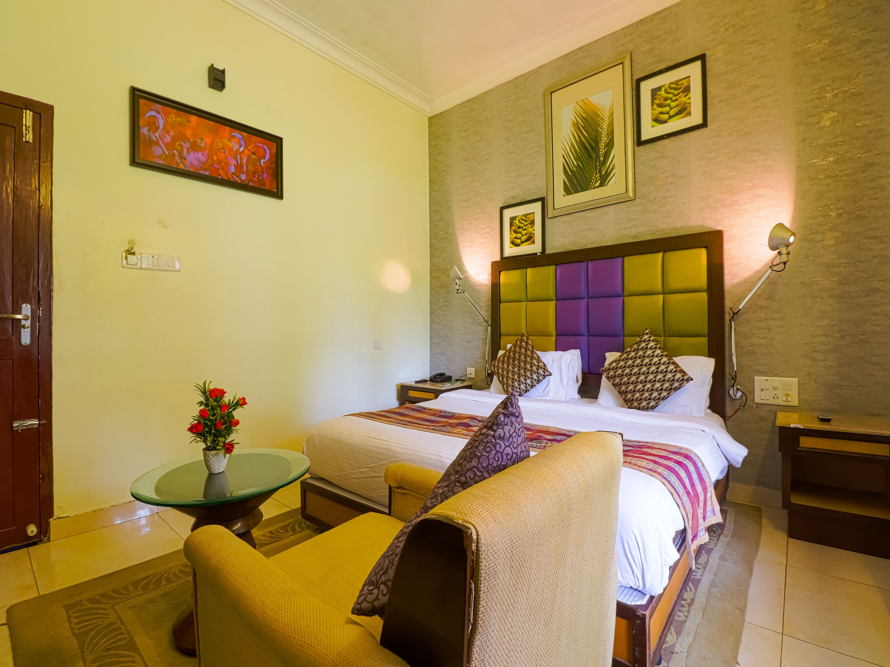 Habitación de hotel con una cama, una zona de estar con una silla, una mesita con un florero y una pared decorada con cuadros.