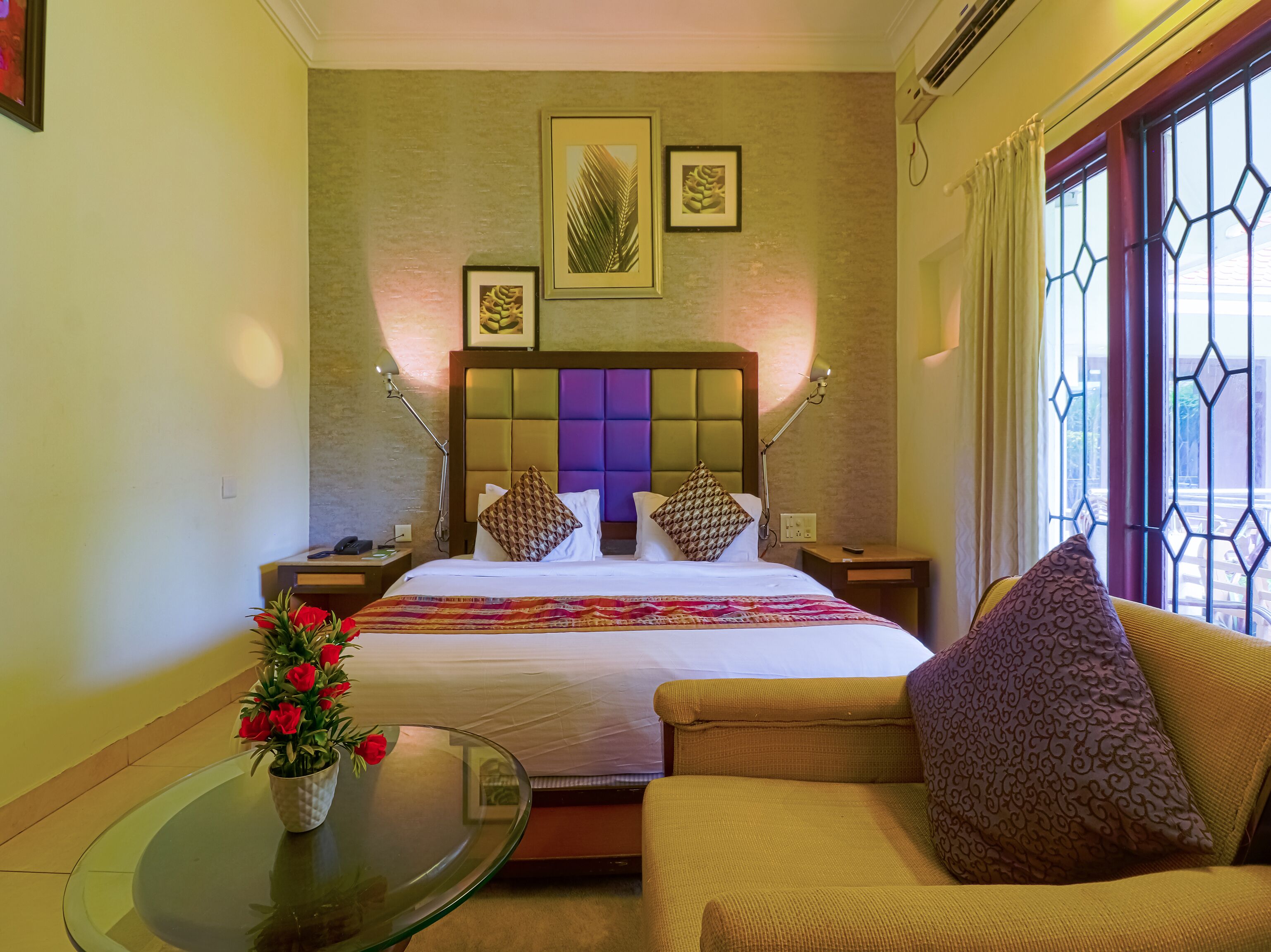 A hotel room with a bed, a chair, a glass table with a vase of flowers, and a window with decorative metal bars.