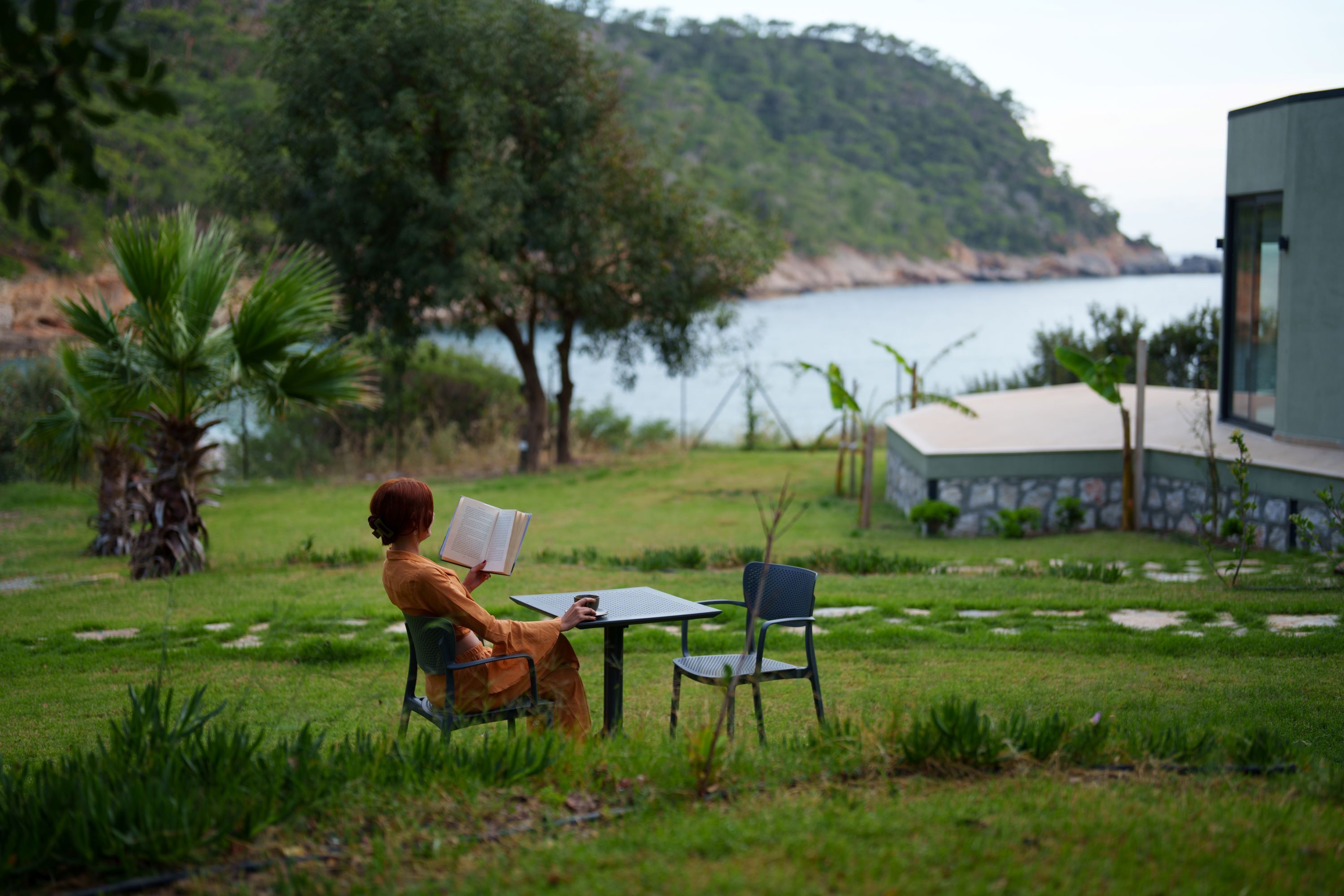 Una persona leyendo en una mesa en un espacio al aire libre con césped, con vistas a un cuerpo de agua y colinas al fondo.