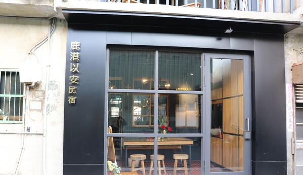 A building with a black facade and a large window. There is a sign with Chinese characters on the left side. Inside, there is a wooden table with stools and a vase of flowers. - LuKang Yian (Lukang)