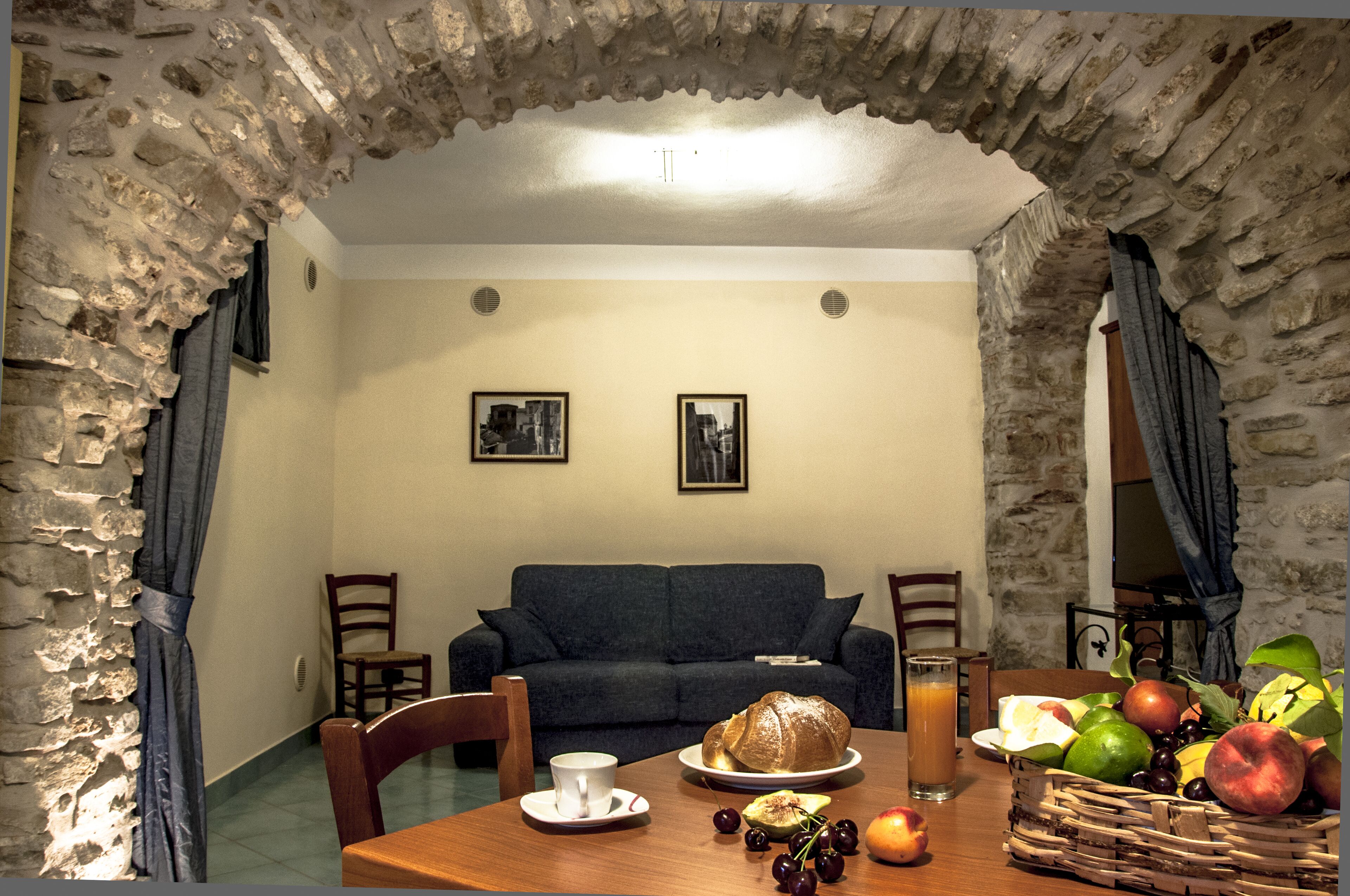A dining area with a stone archway, a sofa, and a table set with a basket of fruit, a croissant, and a glass of orange juice.