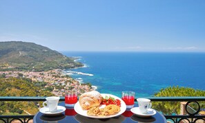 A table with a croissant, cookies, and a plate of red flowers, with two cups of red drink and a white cup, overlooking a coastal town and the sea. - Residenza Tamara (Castellabate)
