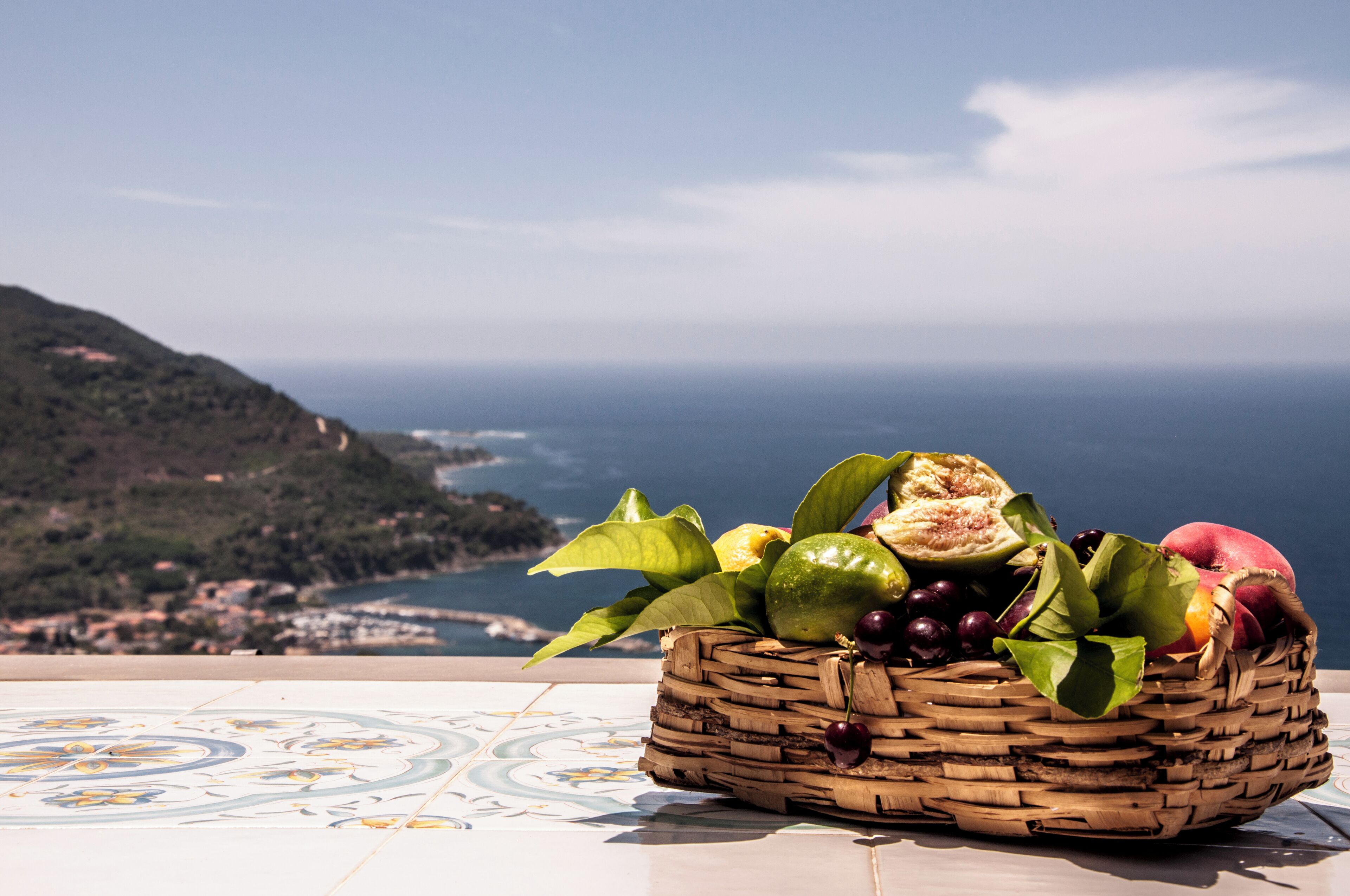 A wicker basket with fruits and vegetables on a tiled floor with a scenic view of the sea and hills.