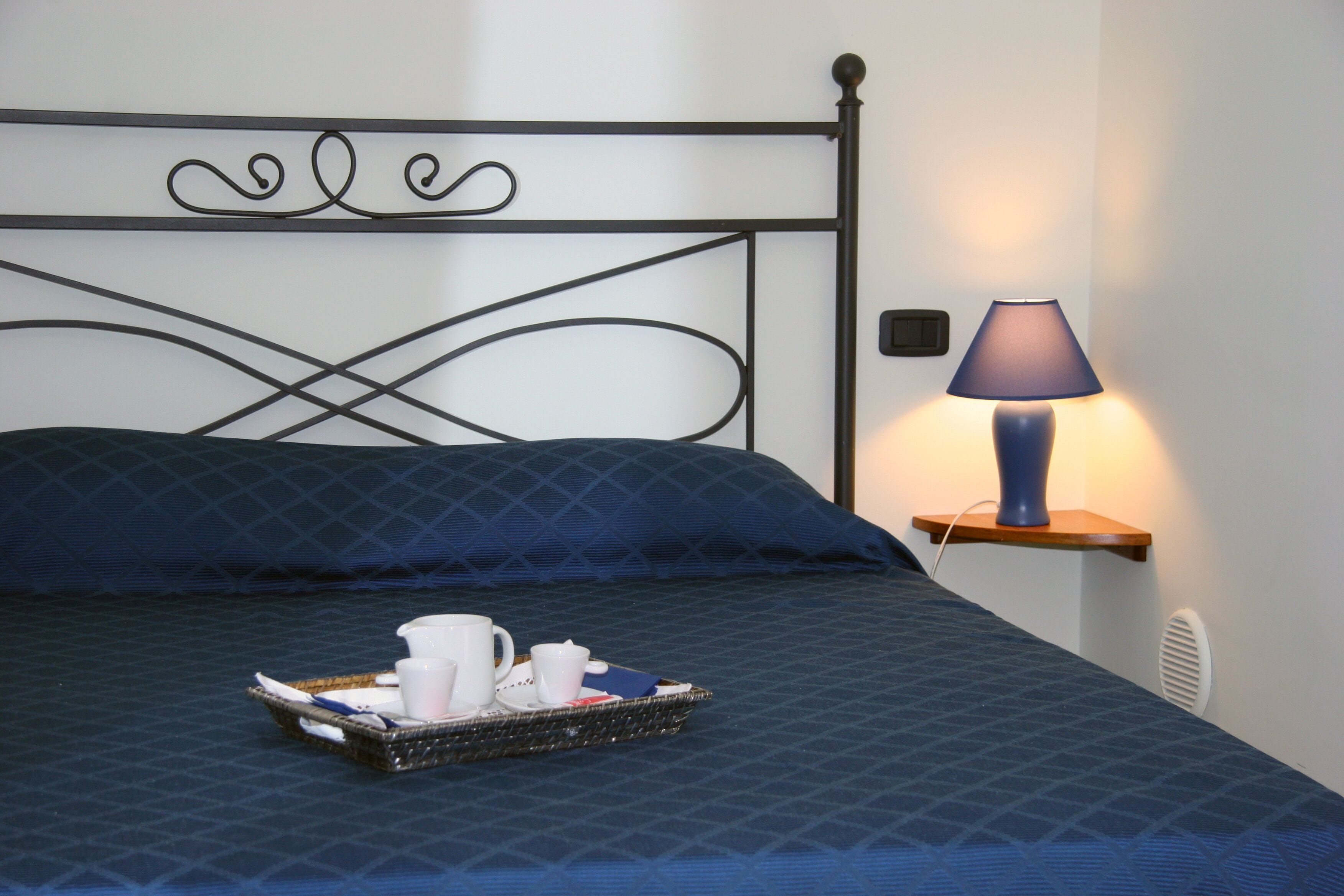 A neatly made bed with a blue bedspread, a tray with cups and a book, and a lamp on a bedside table.