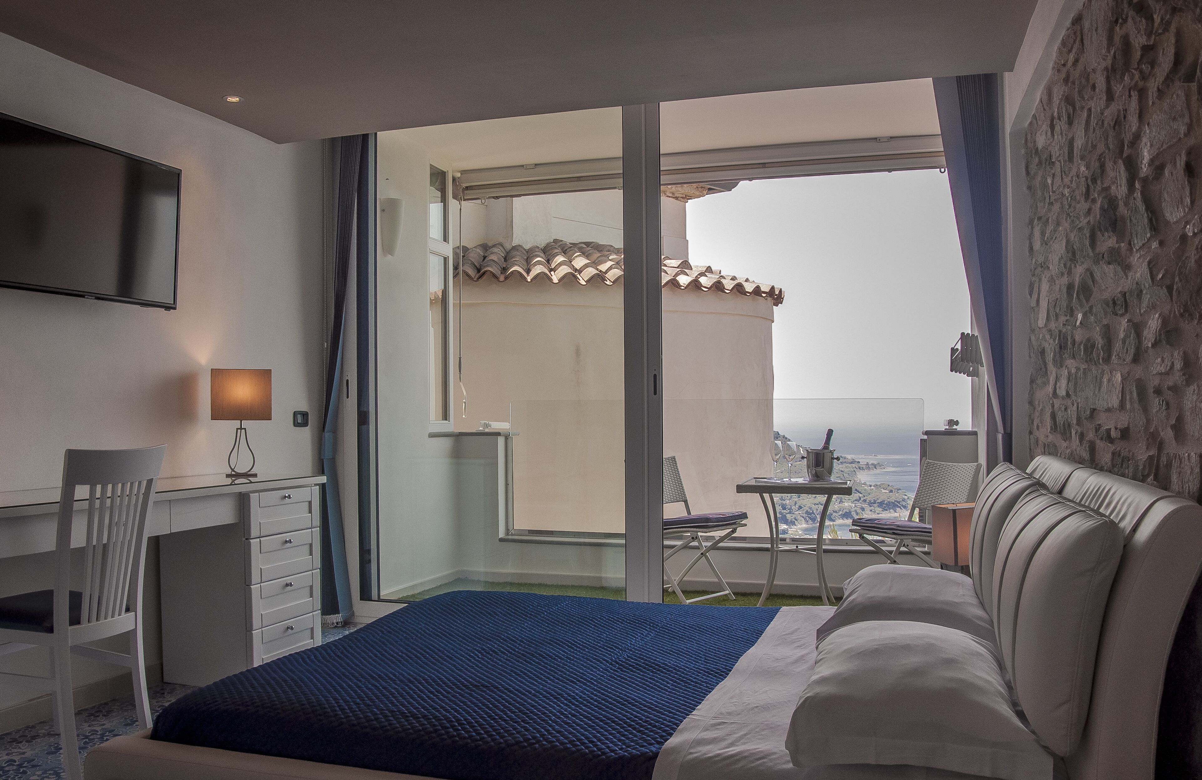 A hotel room with a bed, desk, chair, and a balcony with a view.