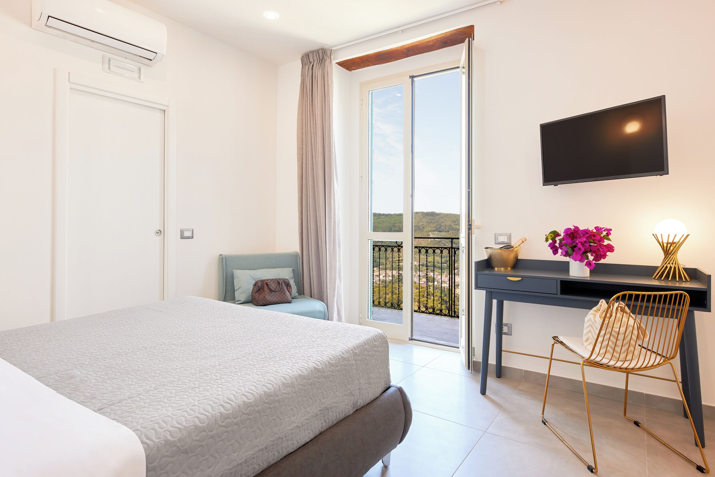 A bedroom with a bed, a desk with a chair, a TV, and a balcony with a view.