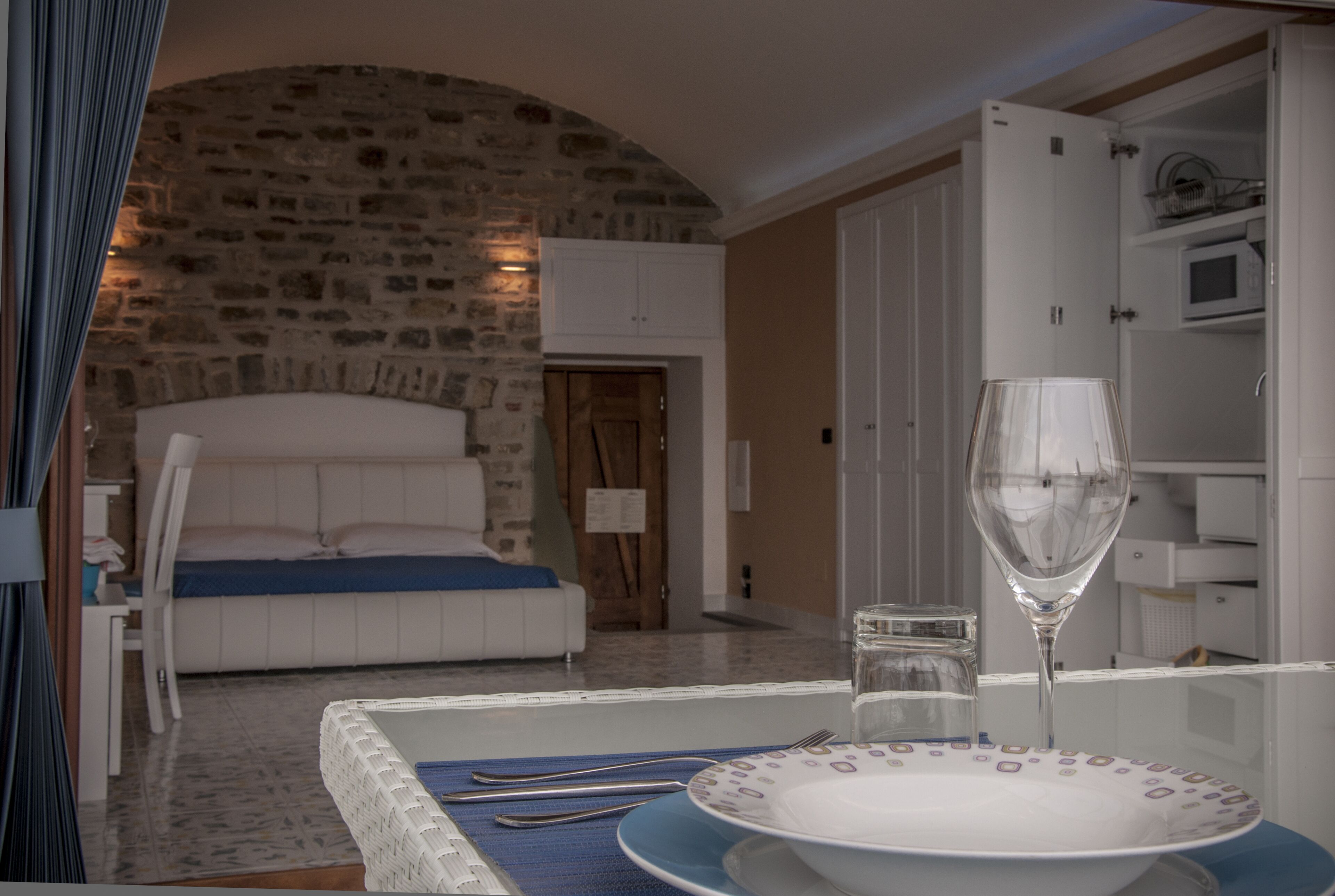 Un dormitorio con pared de piedra, una cama, una mesita de noche con un vaso y un comedor con una mesa lista para dos.