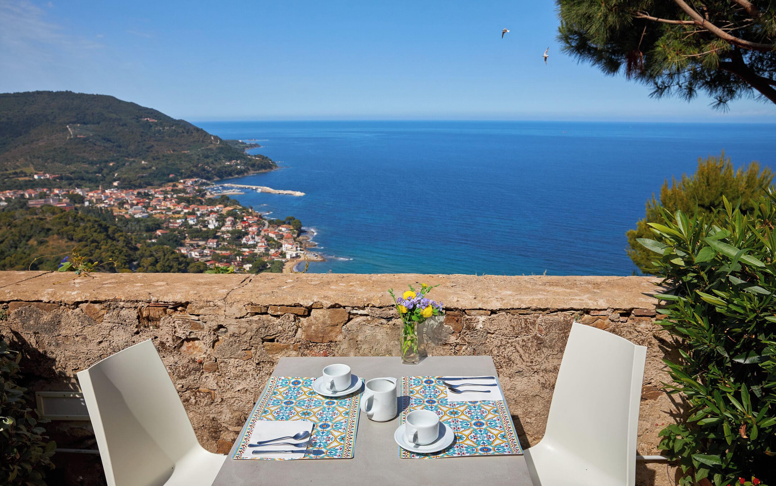 A table set with a vase of flowers, two cups, and a plate, overlooking a coastal town and the sea.