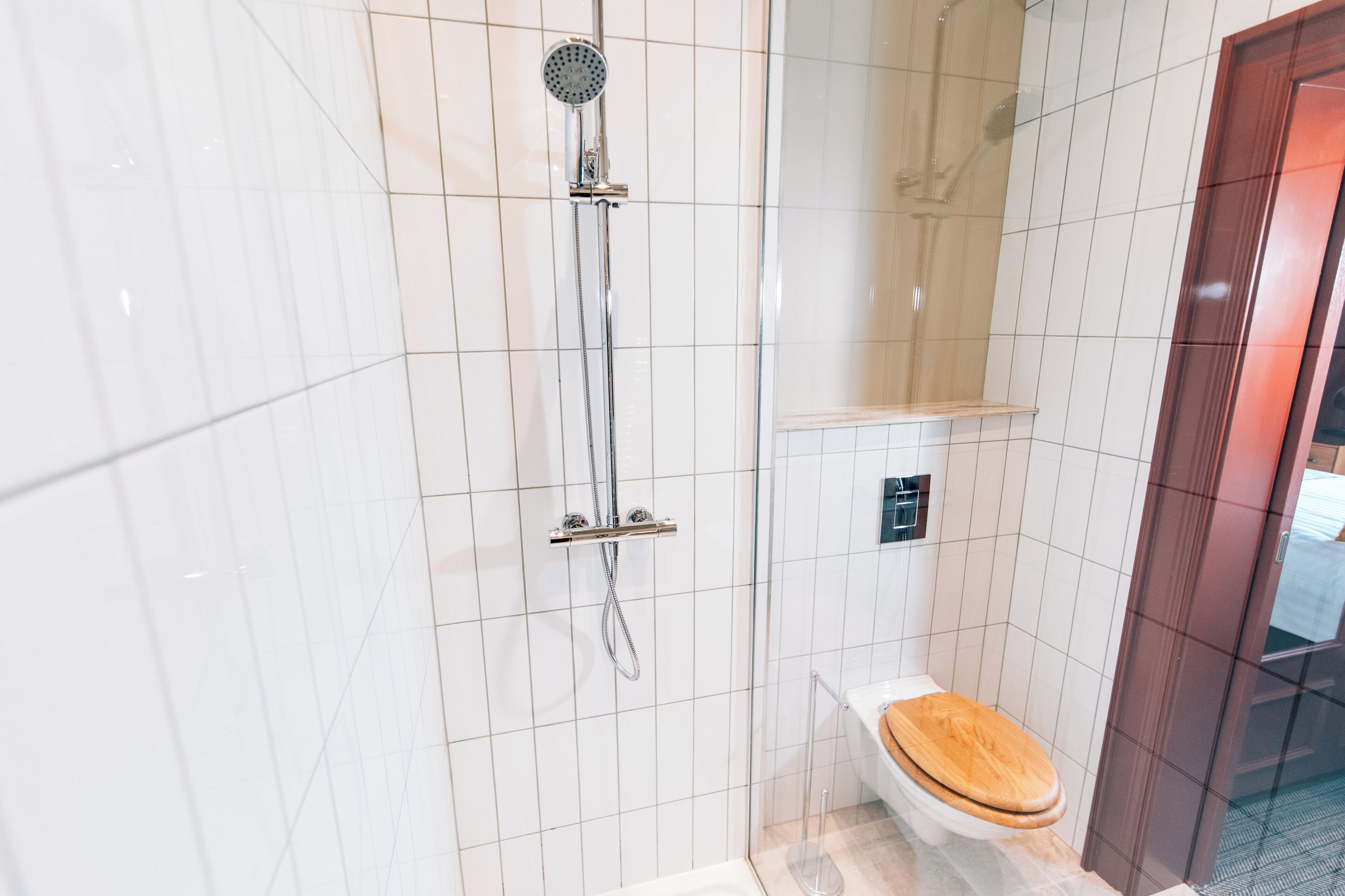 Un baño moderno con paredes de azulejos blancos, una ducha con cabezal de mano, un inodoro con asiento de madera y una mampara de vidrio.