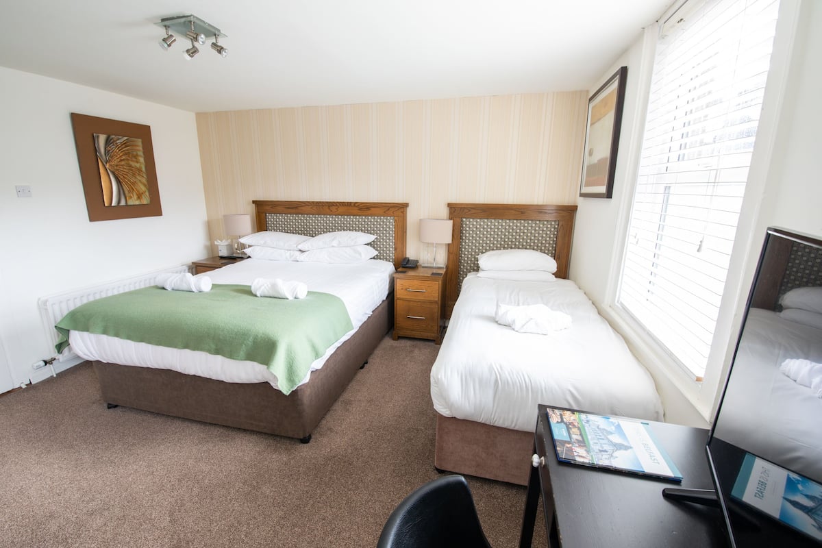 Une chambre d’hôtel avec deux lits, un bureau, une chaise, une lampe, un tableau encadré et une fenêtre avec des stores.