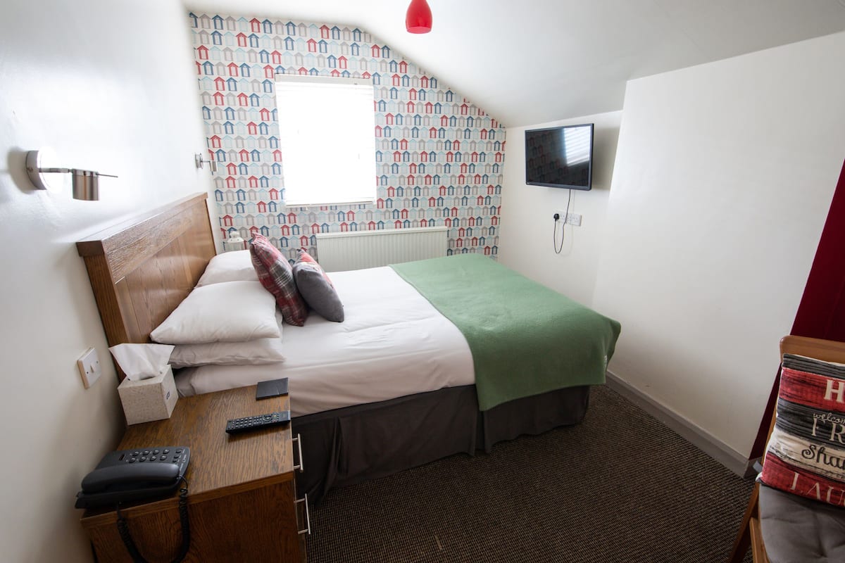 Une chambre d’hôtel comprenant un lit, une table de chevet avec un téléphone et une télécommande, un téléviseur fixé au mur et un papier peint à motifs.