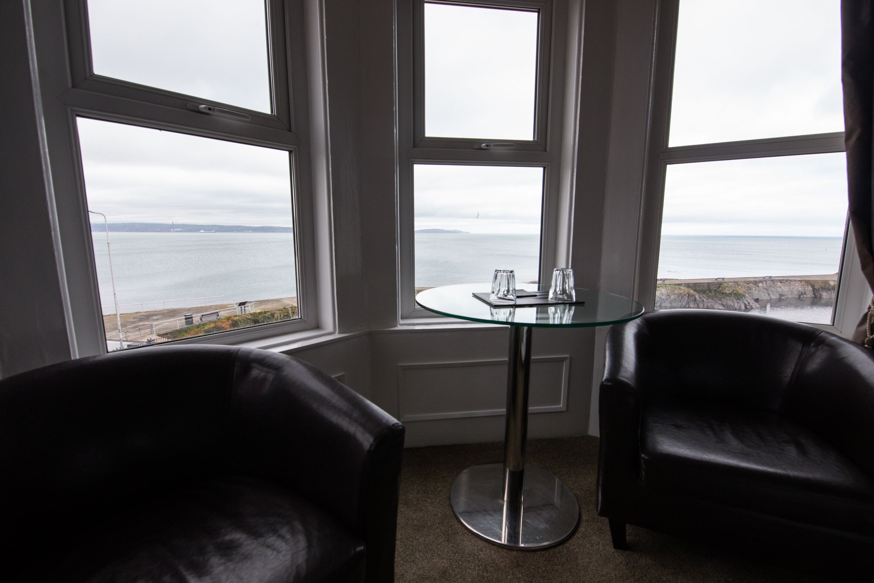 Una habitación con una mesa de vidrio, dos sillas de cuero negro y grandes ventanales con vistas al mar.