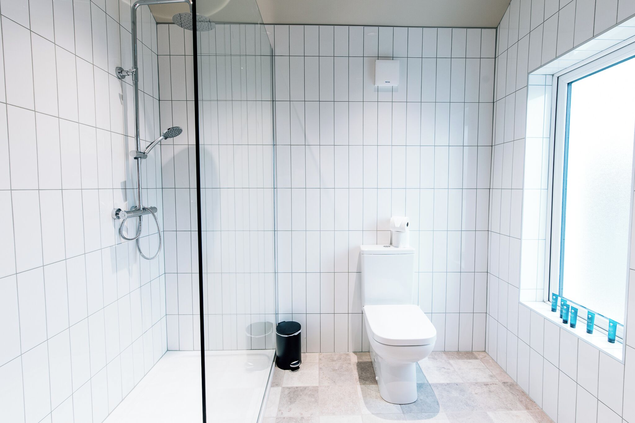 A modern bathroom with white tiles, a glass shower enclosure, a toilet, and a window with a view.