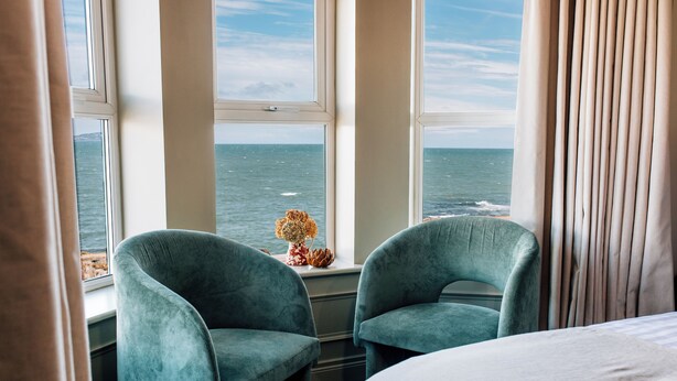 A room with two green armchairs by a window overlooking the sea, a bed, and a decorative vase with flowers on the windowsill. - The Nines, Bangor Marina (Bangor)