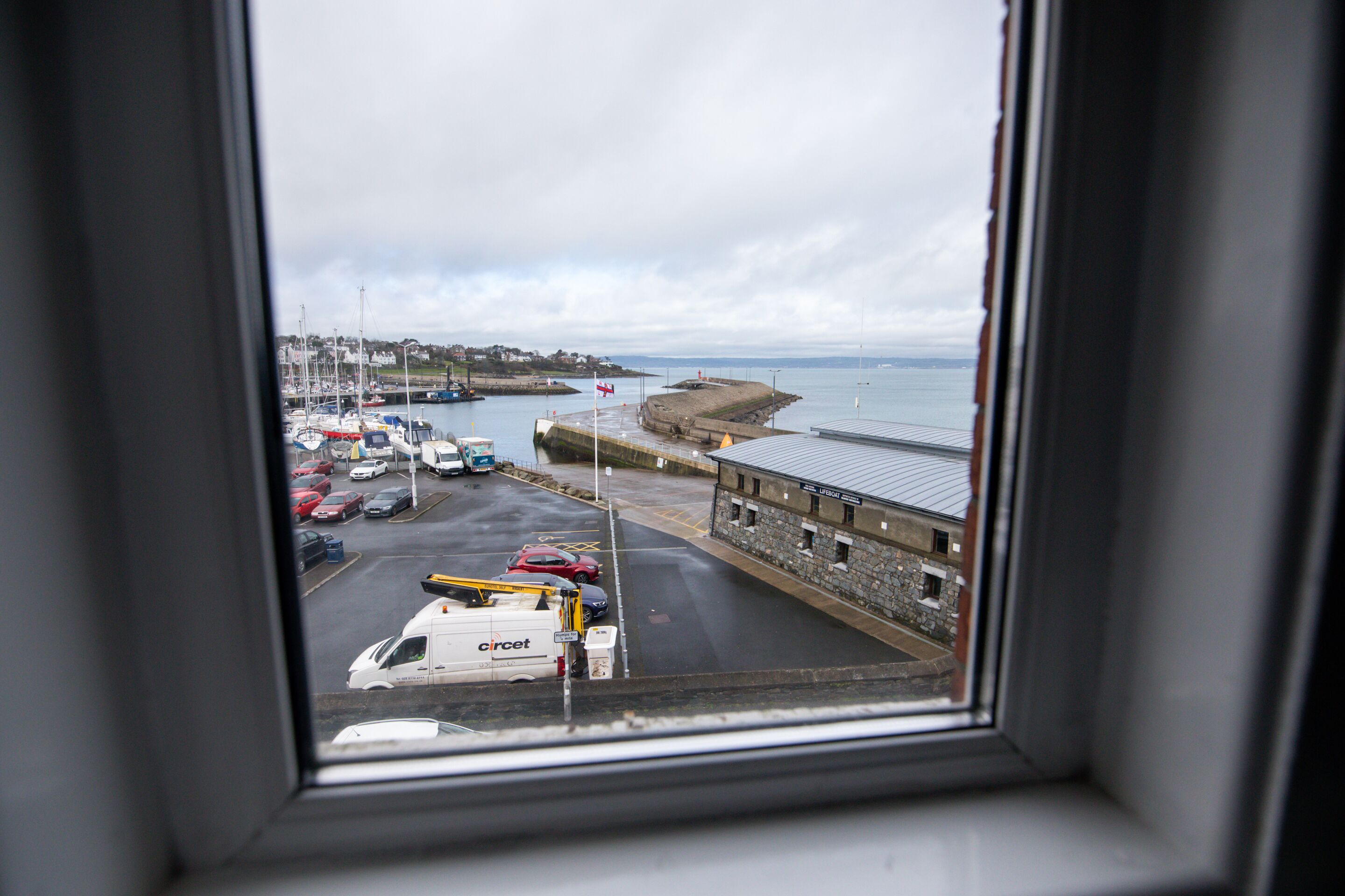 A harbor view from a window, including parked vehicles, a building, and a flag.