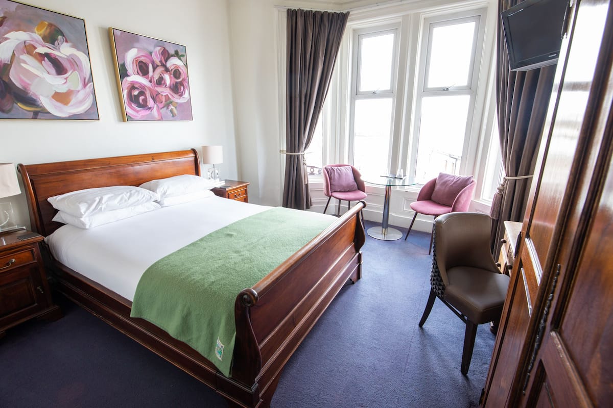 Une chambre d’hôtel avec un lit en bois, un plaid vert, deux tableaux, une télévision, un bureau et une chaise.
