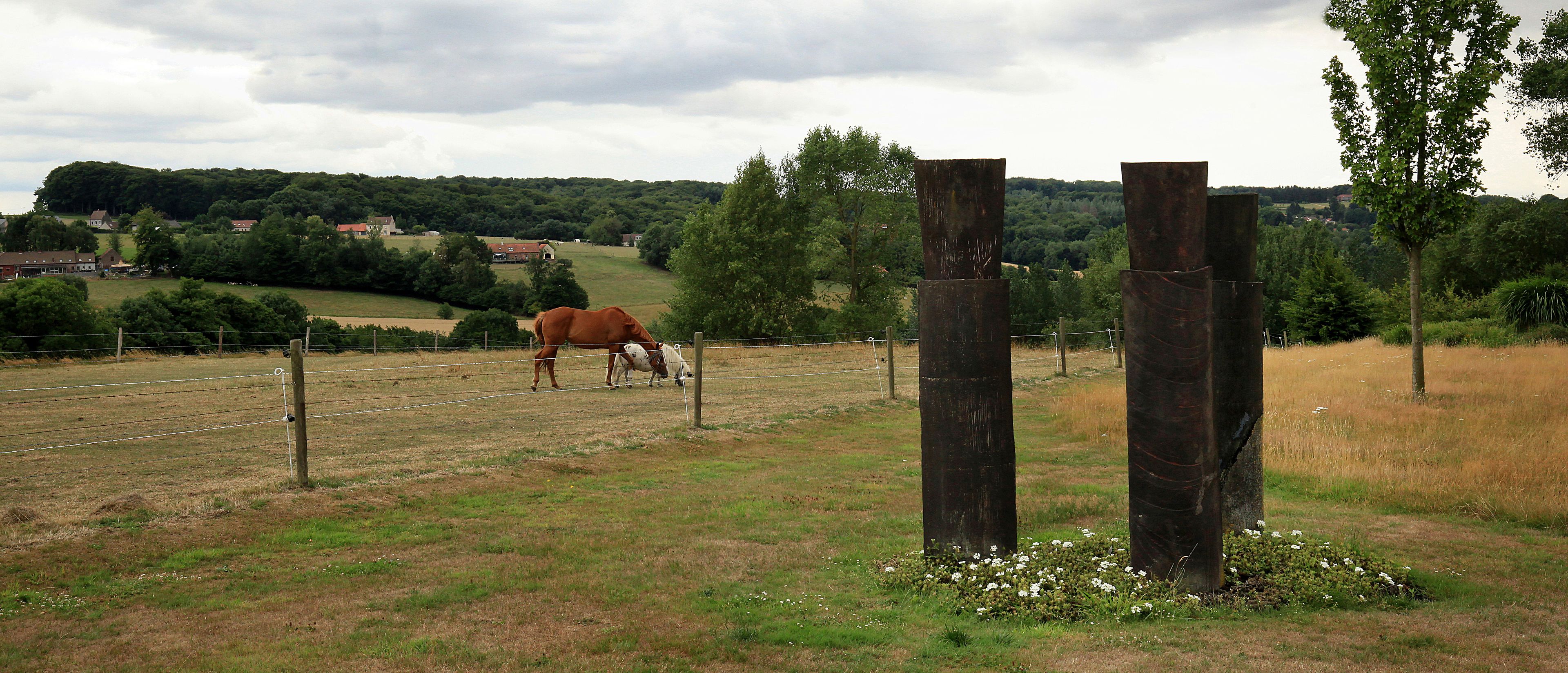 A rural landscape with a horse grazing near a fence, surrounded by greenery and a few buildings in the distance.